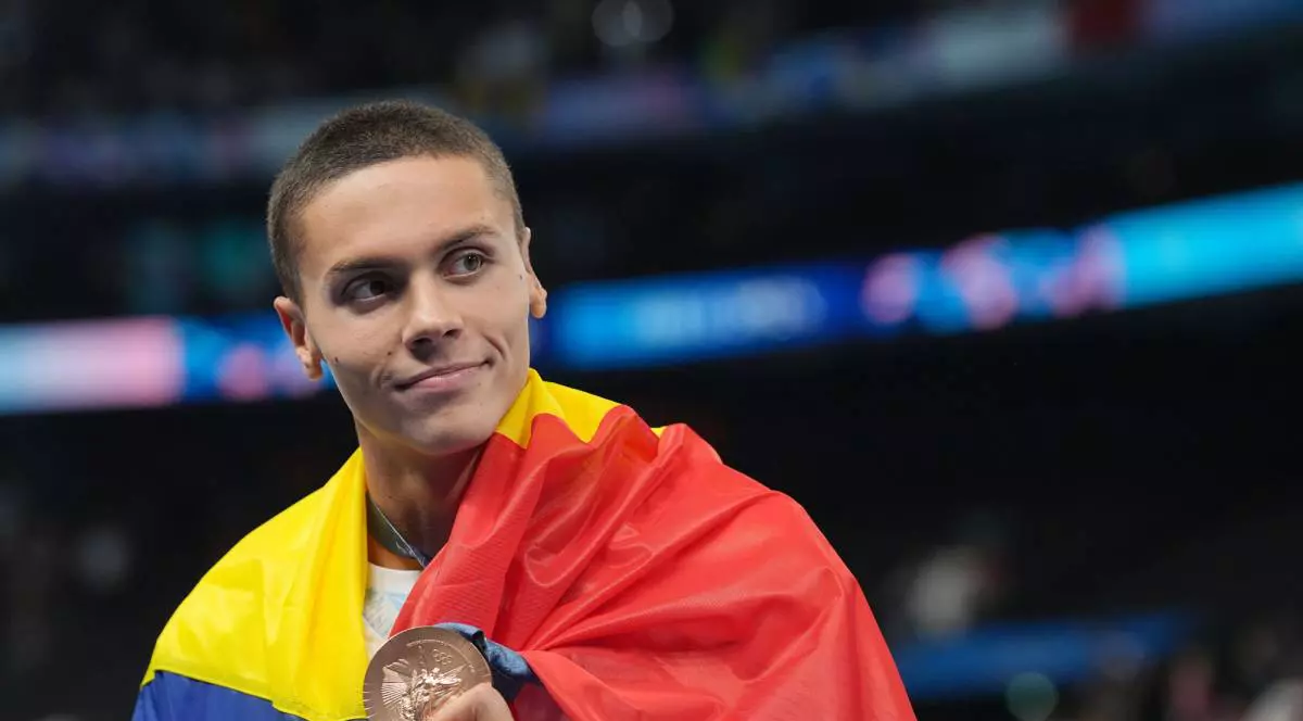 David Popovici cu steagul României pe spate, în timpul unei competiții sportive, zâmbește