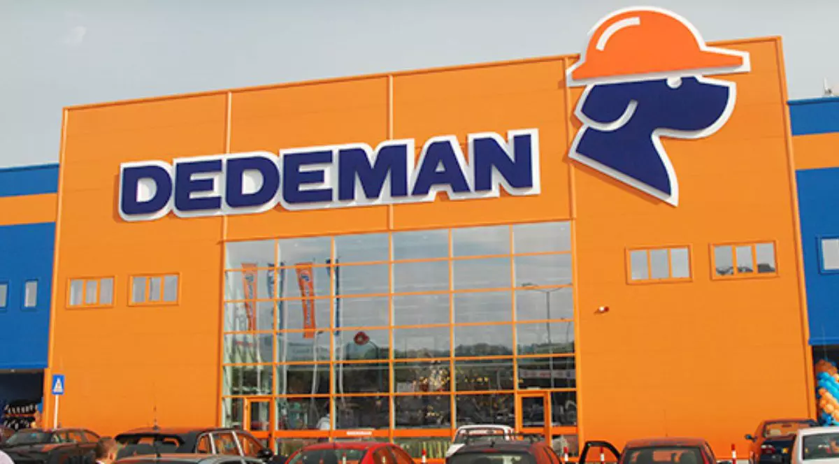 fațada unui magazin de bricolaj Dedeman, vopsit în culorile caracteristice (albastru șu portocaliu), cu logo-ul vizibil. Logo-ul este format din silueta unui cap de cățel colorată în albastru, din profil, cu o cască de protecție portocaliu în cap