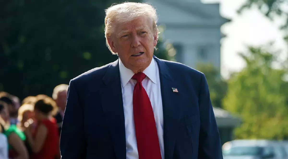Donald Trump, îmbrăcat la costum, cu o cravată vișinie, se plimbă în aer liber, în spatele său se vede un grup de oameni