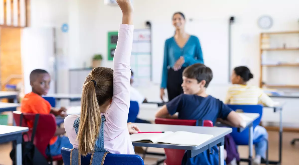 Imagine cu mai mulți elevi care stau în bănci, iar în fața lor se află profesoara. O elevă are o mână ridicată
