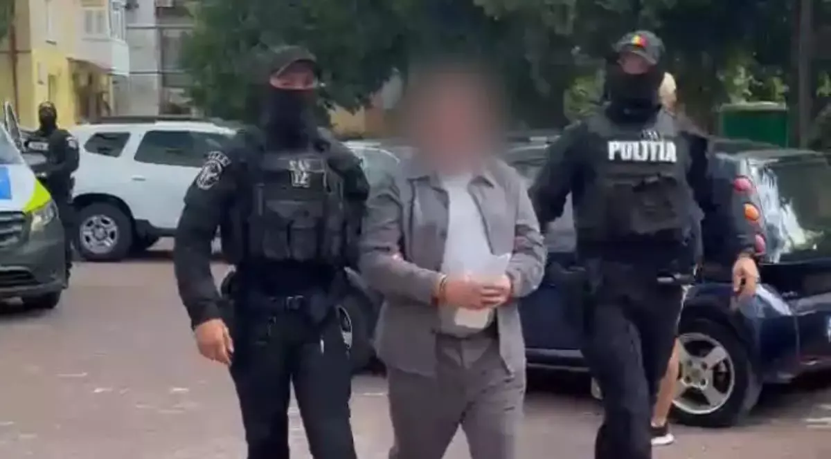 Un bărbat acuzat că a ucis la comandă un tânăr în centrul Berlinului în 2019, a fost arestat la București, iulie 2025. In imagine suspectul este cu fața blurată, cătușe la mâini, insoțit de 2 mascați, intr-o parcare in aer liber