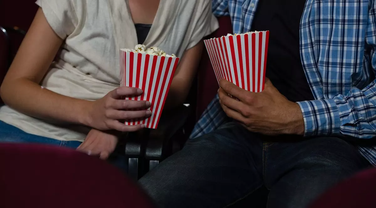 Imagine cu o femeie (în stânga) și un bărbat (în dreapta) în timp ce se află la cinema. Țin în mâini recipiente cu popocorn. Femeia poartă o bluză de culoare deschisă, iar bărbatul o cămașă albastră în carouri și o bluză neagră