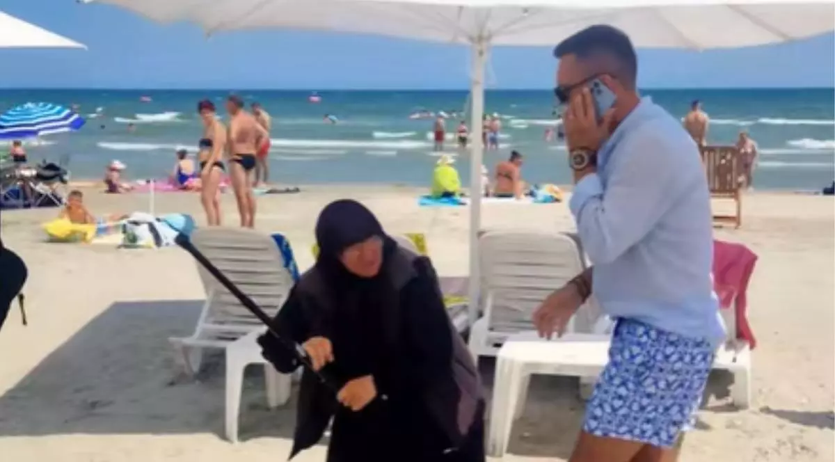 Imagine cu femeia din Republica Moldova (stânga) în timp ce ridică bastonul pentru a-l lovi pe administratorul plajei din Mamaia (dreapta). Femeia este îmbrăcată în negru și are capul acoperit cu o eșarfă, iar bărbatul vorbește la telefon și poartă o cămașă în culoarea albastru deschis și pantaloni scurți cu alb și albastru
