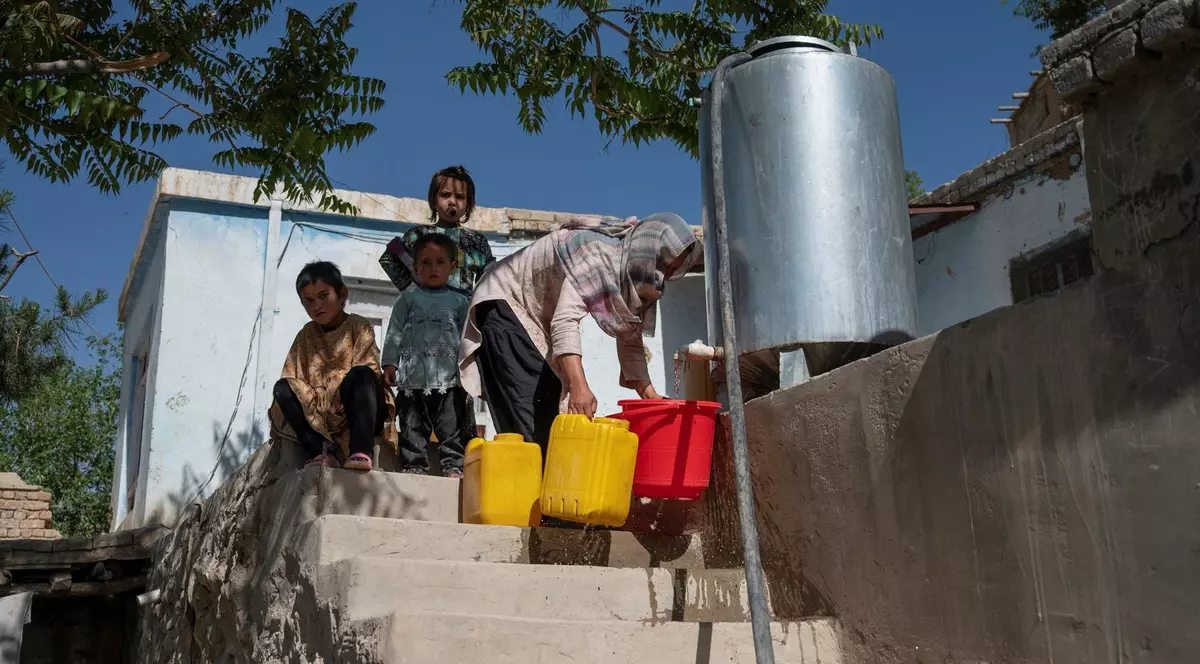 Imagine cu o femeie din Kabul care umple 3 recipiente cu apă dintr-o cisternă. Este însoțită de trei copii.