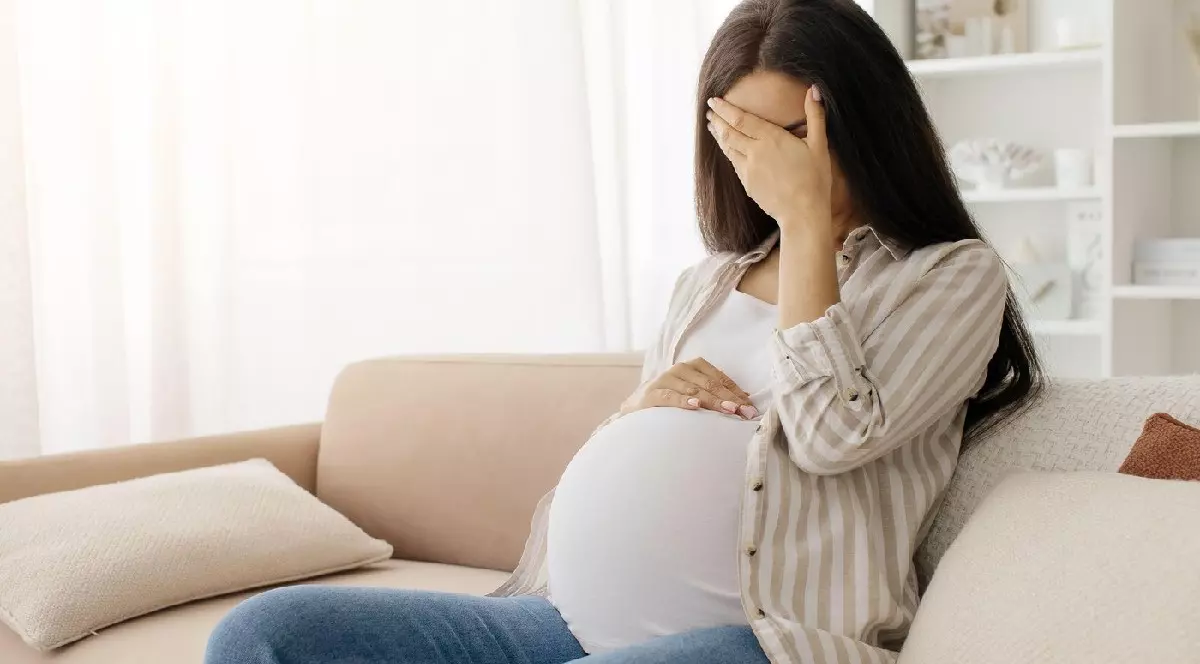 O femeie insarcinată, îmbrăcată cu maieu alb, blugi și o cămașă cu dungi albe și bej, stă așezată pe o canapea bej. Își acoperă fața cu o mână