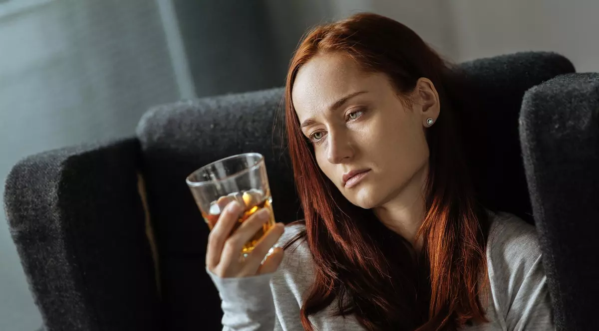 Imagine cu o femeie care stă într-un fotoliu negru și care ține în mână un pahar. Aceasta privește paharul și poartă o bluză gri