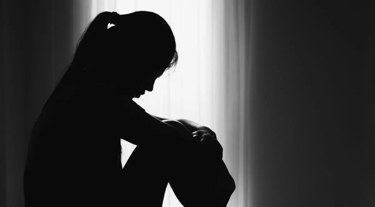 Imagine alb-negru cu silueta unei femei asezata, cu spatele la un perete, capul plecat, genunchii strânși la piept, mainile pe genunchi, ilustrand conceptul de singuratate