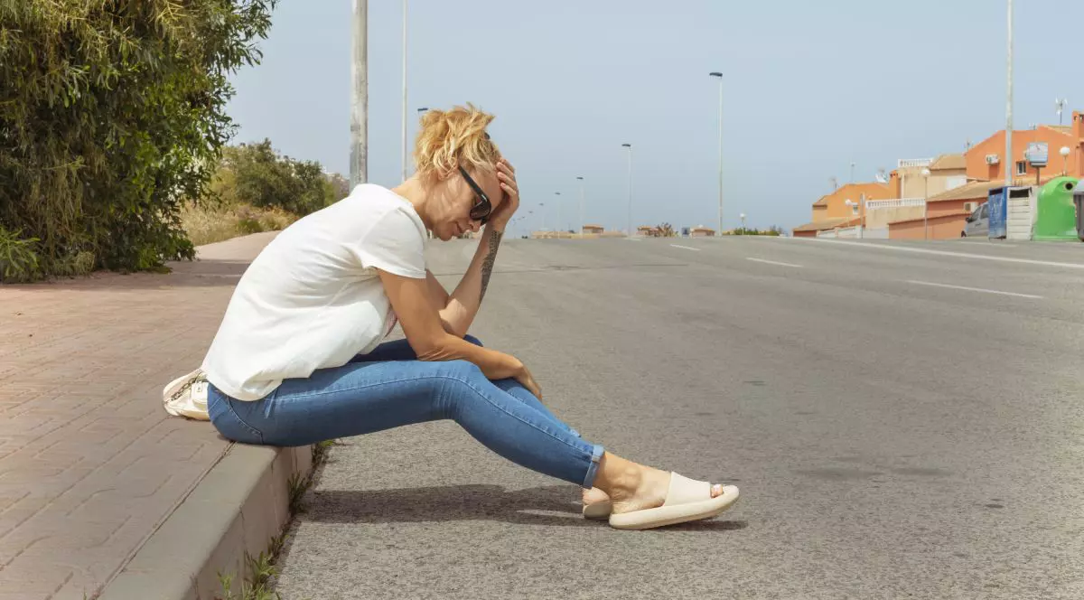 Femeie îmbrăcată cu o bluză albă, blugi albaștri și papuci crem, supărată, în timp ce stă pe marginea unui trotuar, lăngă o stradă goală