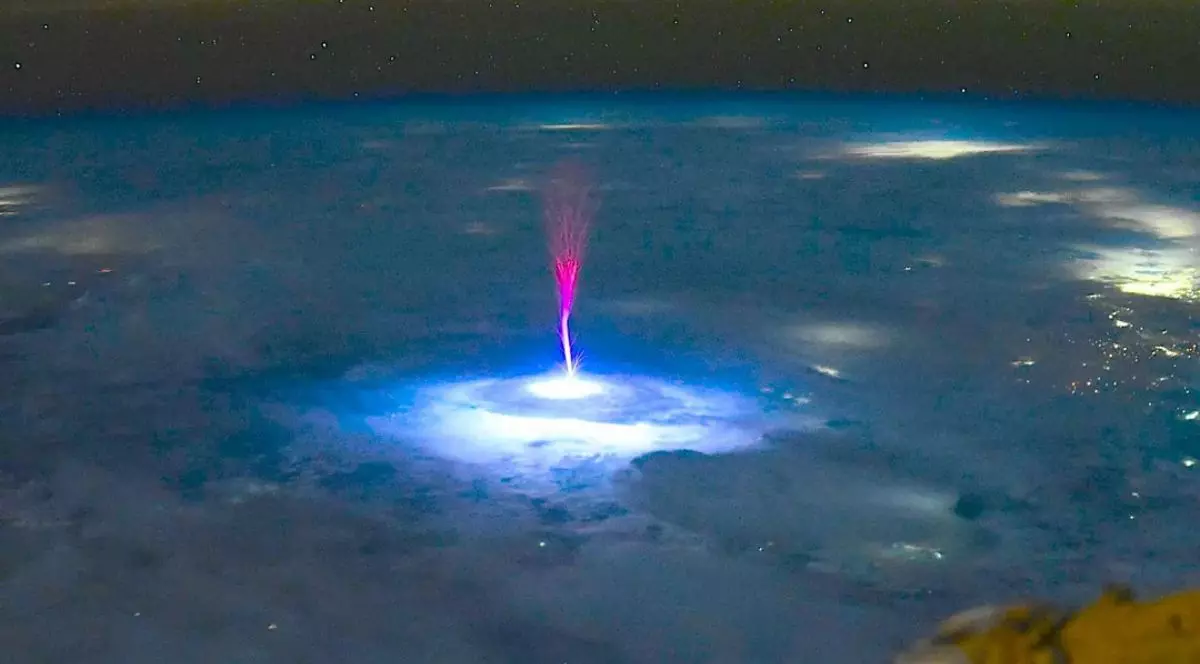 explozie de lumină albastră intensă, ca o pată circulară, care luminează norii de sub - în mijlocul acestei zone luminoase pornește un jet vertical de lumină roșie-violet, care se ridică în sus spre cer