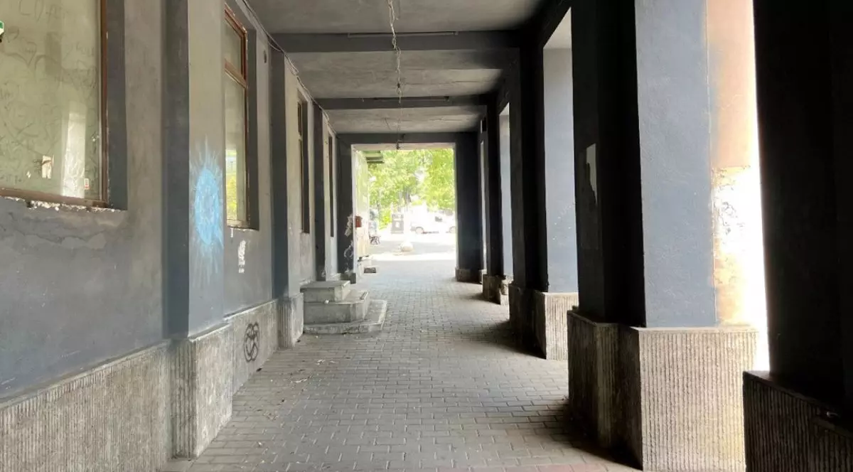 coridor lung, acoperit, cu tavan drept și mai mulți stâlpi masivi de susținere amplasați de-a lungul marginii din dreapta. Podeaua este pavată cu dale dreptunghiulare de culoare gri. În partea stângă se află un zid de culoare gri-închis, în care sunt încadrate ferestre mari, cu rame maro, poziționate la intervale regulate. Pe același perete sunt vizibile urme de graffiti sau pete de vopsea, precum și câteva inscripții sau mâzgălituri.