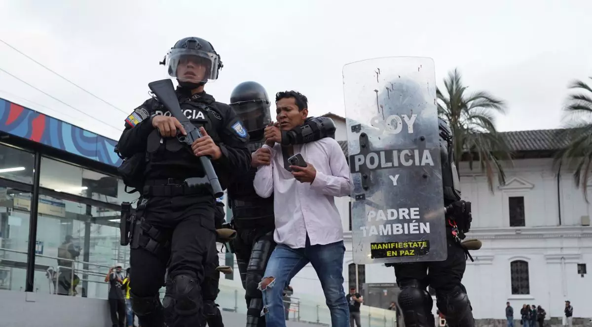 Trei polițiști cu căști, scuturi și arme automate escortează un bărbat care ține în mână un telefon mobil, în Quito, Ecuador, noiembrie 2024
