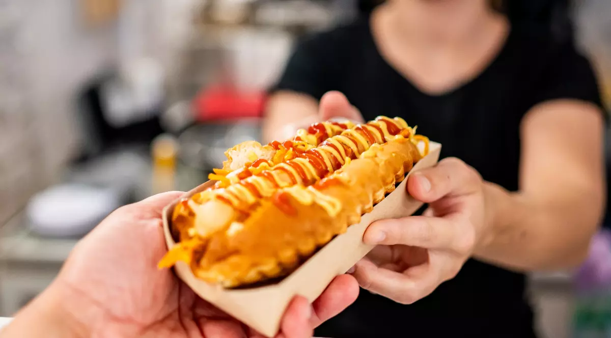 O femeie care ține în mână un hot-dog cu muștar și ketchup pe care-l oferă unui om