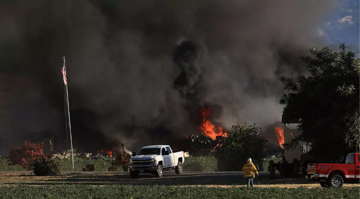 incendiu de vegetație care degază fum negru și gros. în partea de jos a imaginii se vede o pajiște verde, cu un pompier îmbrăcat în echipament galben, și două mașini pick-up mari. în spatele mașinilor se văd flăcări de culoare roșu aprins. în stânga imaginii se observă un catarg pe care este prins steagul SUA, care stă culcat în jos