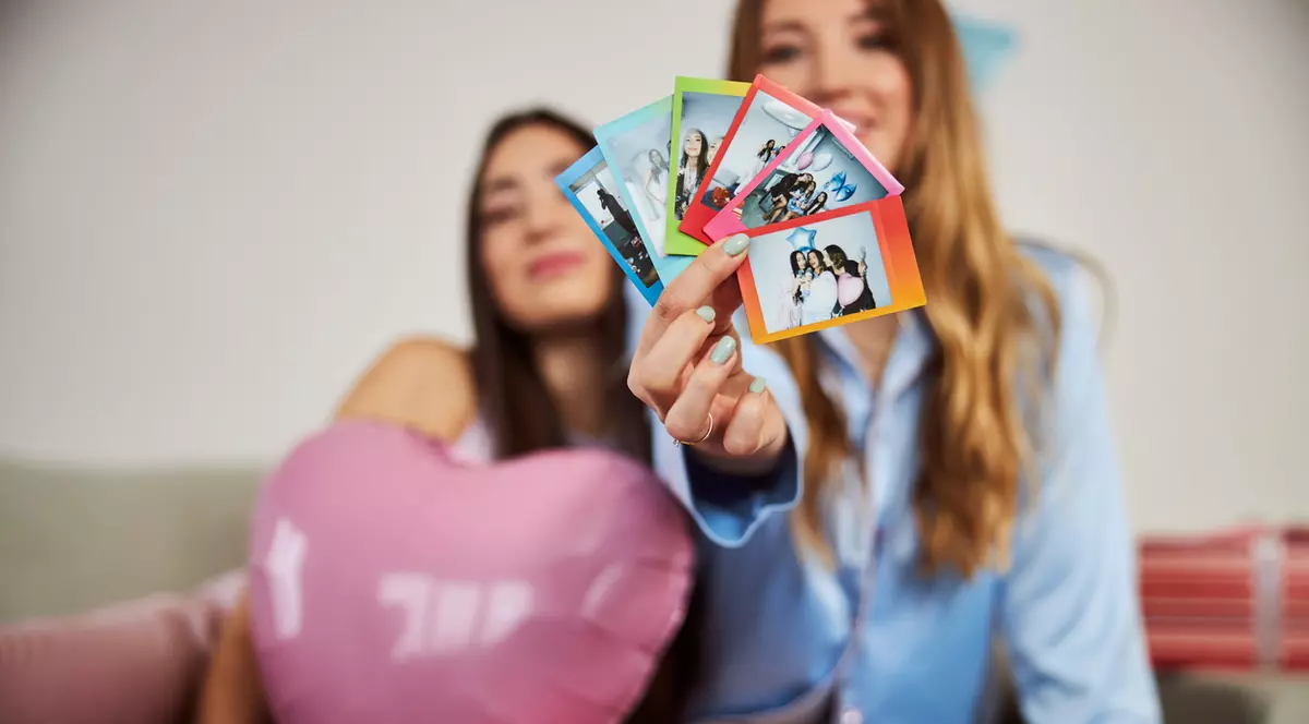 Doua fete, una bruneta si una blonda. Cea bruneta este in stanga si tine in brate un balon roz in forma de inima, cea blonda sta in drepata si tine in mana poze instant