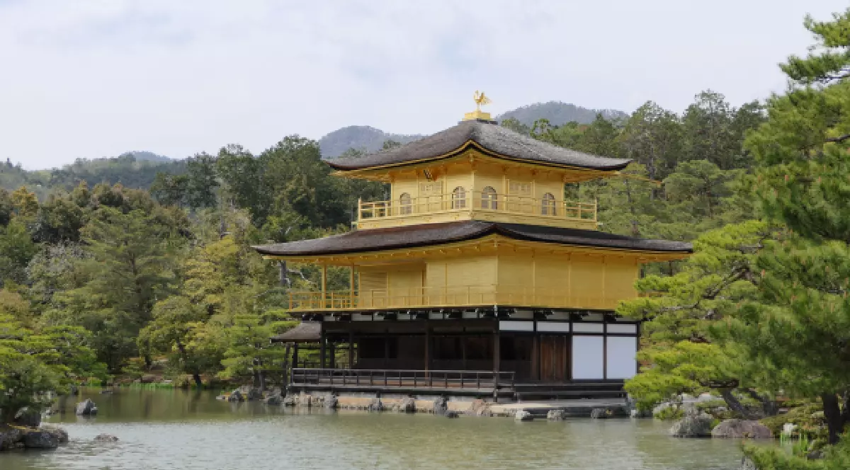 O clădire religioasă aurie tradițională japoneză se reflectă în apa liniștită a unui iaz, înconjurată de pini și dealuri verzi.