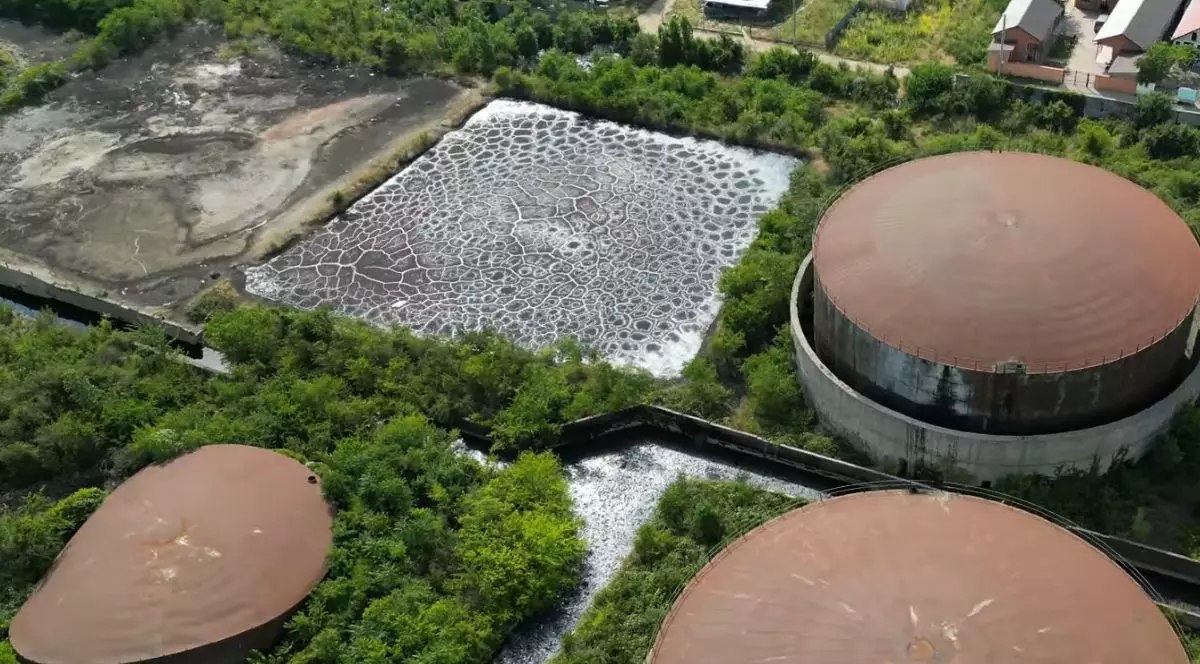 Fotografie realizată dintr-un unghi aerian și surprinde o zonă extinsă, în mare parte acoperită de vegetație verde. Sunt vizibile trei structuri mari, de formă circulară, cu acoperiș metalic de culoare ruginie, două dintre ele având diametre egale și una cu formă ovală. În partea din stânga sus se poate observa un lac acoperit de ce par a fi pete de ulei