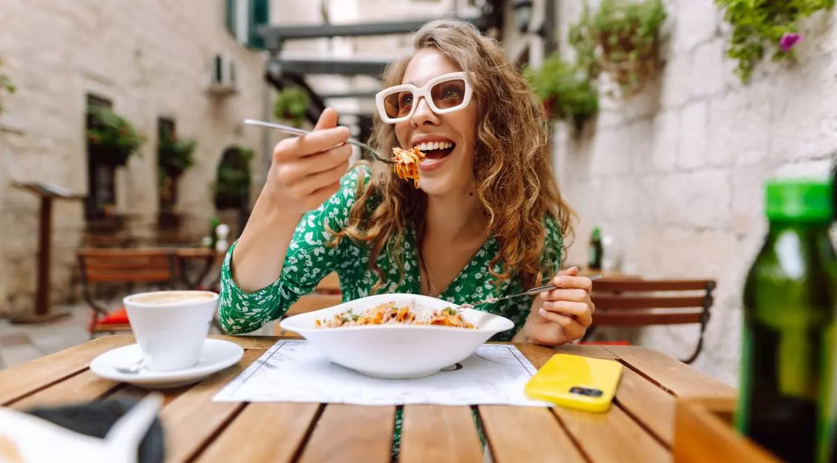 O femeie tânără, cu o bluză verde și ochelari de soare cu rame albe, stă la masă pe terasa unui restaurant și mânâncă paste cu roșii dintr-un castron alb. Are pe masă un telefon galben și o cafea