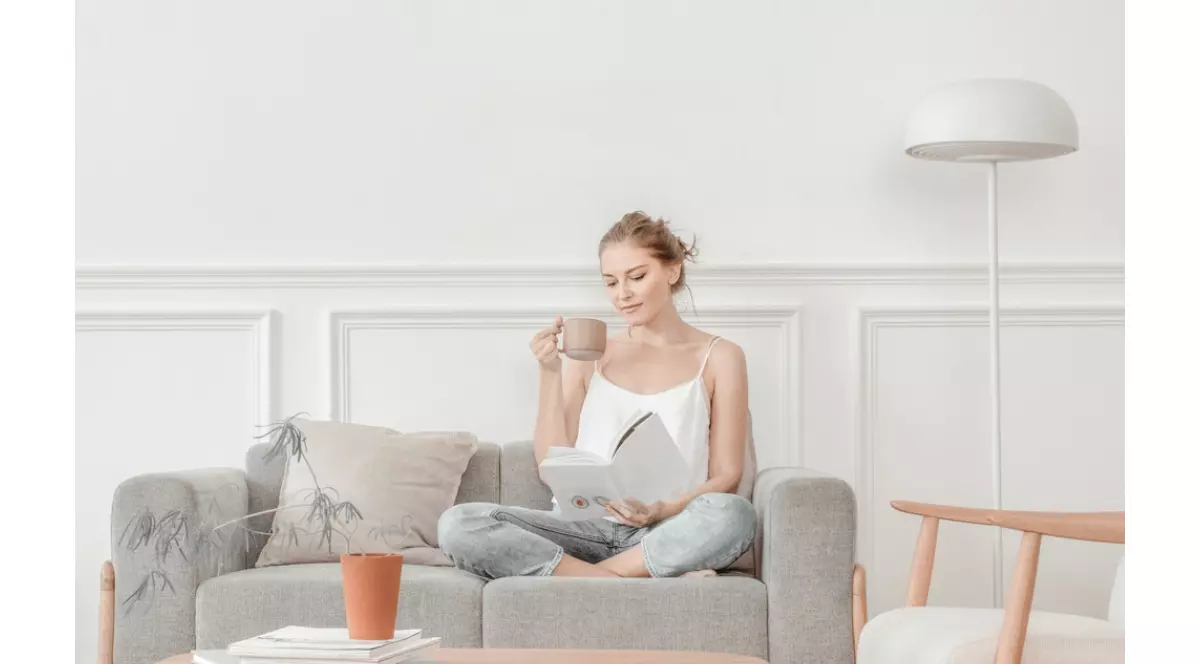 Femeie care sta in sufragerie pe o canapea de 3 persoane gri, se relaxeaza cu o cafea in mana si citind