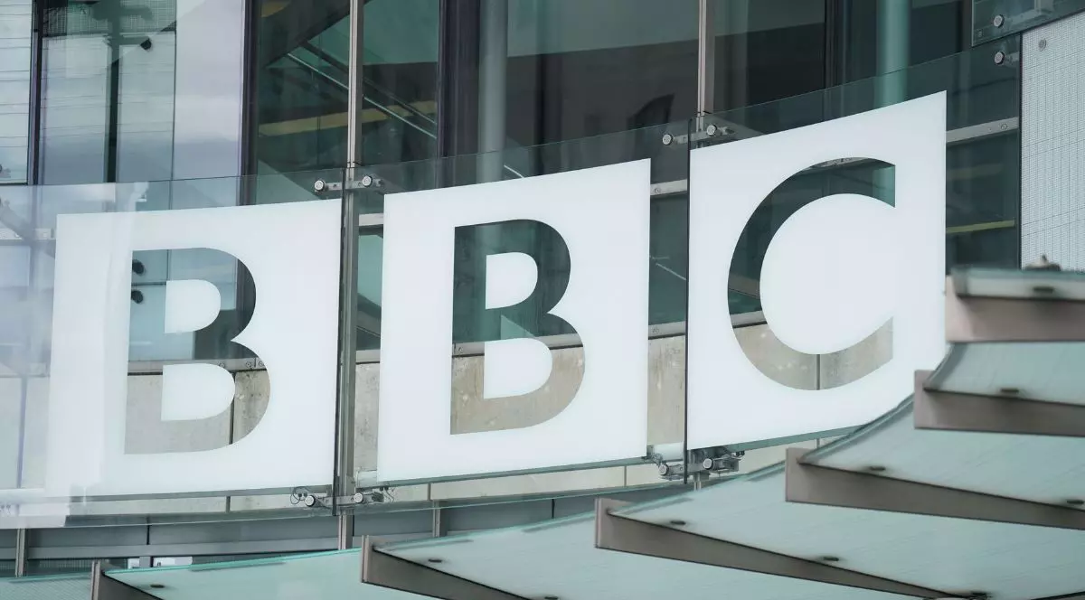 clădire modernă cu fațadă de sticlă. Pe partea frontală, sunt trei panouri albe, montate unul lângă altul, fiecare cu o literă neagră, formatând împreună cuvântul „BBC”. Panourile sunt fixate de o balustradă transparentă cu cleme metalice vizibile. Sub aceste panouri, în prim-plan, se află un acoperiș din sticlă susținut de stâlpi cilindrici gri, încorporați în structura clădirii. Reflecții ale stâlpilor și panourilor apar pe suprafața de sticlă, laolaltă cu contururi vagi ale scărilor și balustradelor din interiorul clădirii