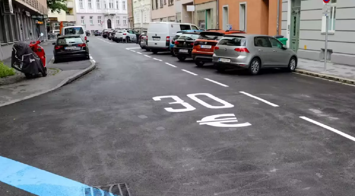 O stradă asfaltată, cu mașini parcate pe partea dreaptă, iar pe carosabil este pictat mare cu alb „30” alături de simbolul euro, lângă marcaje de parcare trasate cu linii albe.