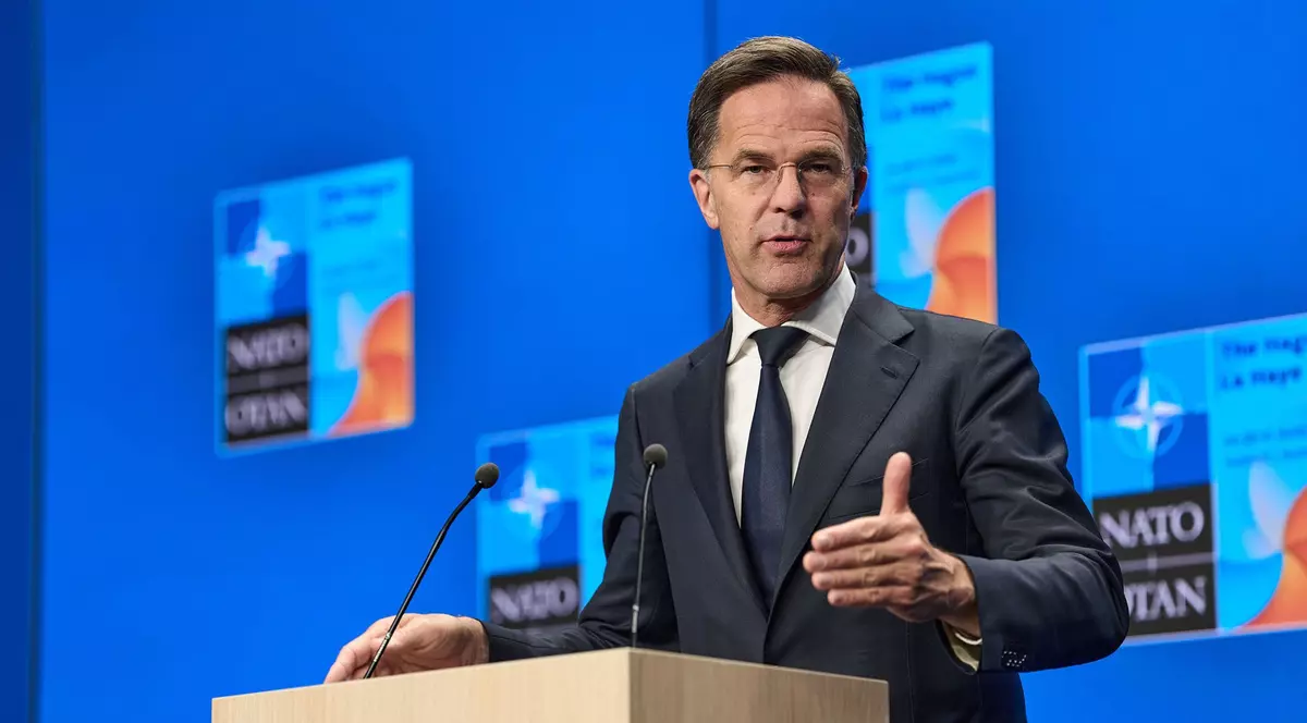 Mark Rutte surprins în timpul unei conferințe de presă la summitul NATO. Acesta are o mână ridicată și poartă un sacou negru, o cămașă albă și o cravată neagră