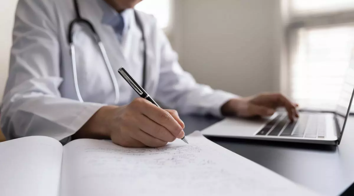 Un medic de familie, în timp ce stă la birou, în fața laptopului și notează datele pacientului asigurat la sănătate.