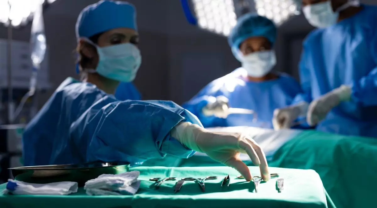 Trei medici în costume sterile albastre, cu masti pe față in timpul unei operații, in afat se vede instrumentarul steril