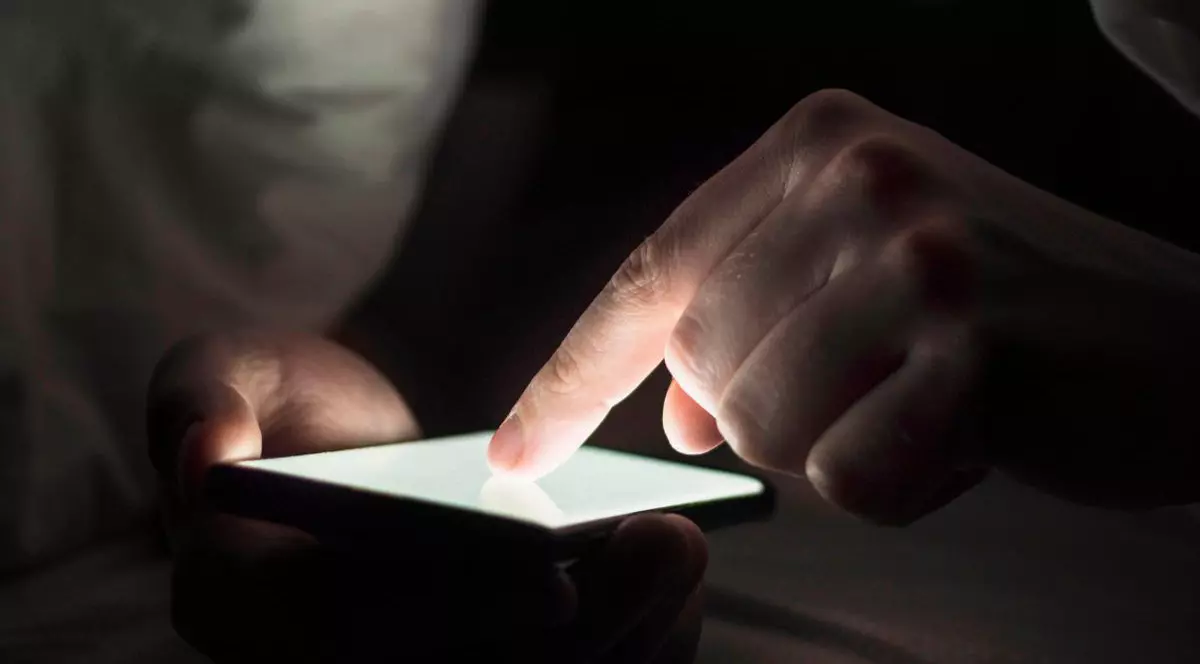 Bărbat care stă pe telefon, ținând degetul arătător lipit de ecranul telefonului mobil care este luminat în alb