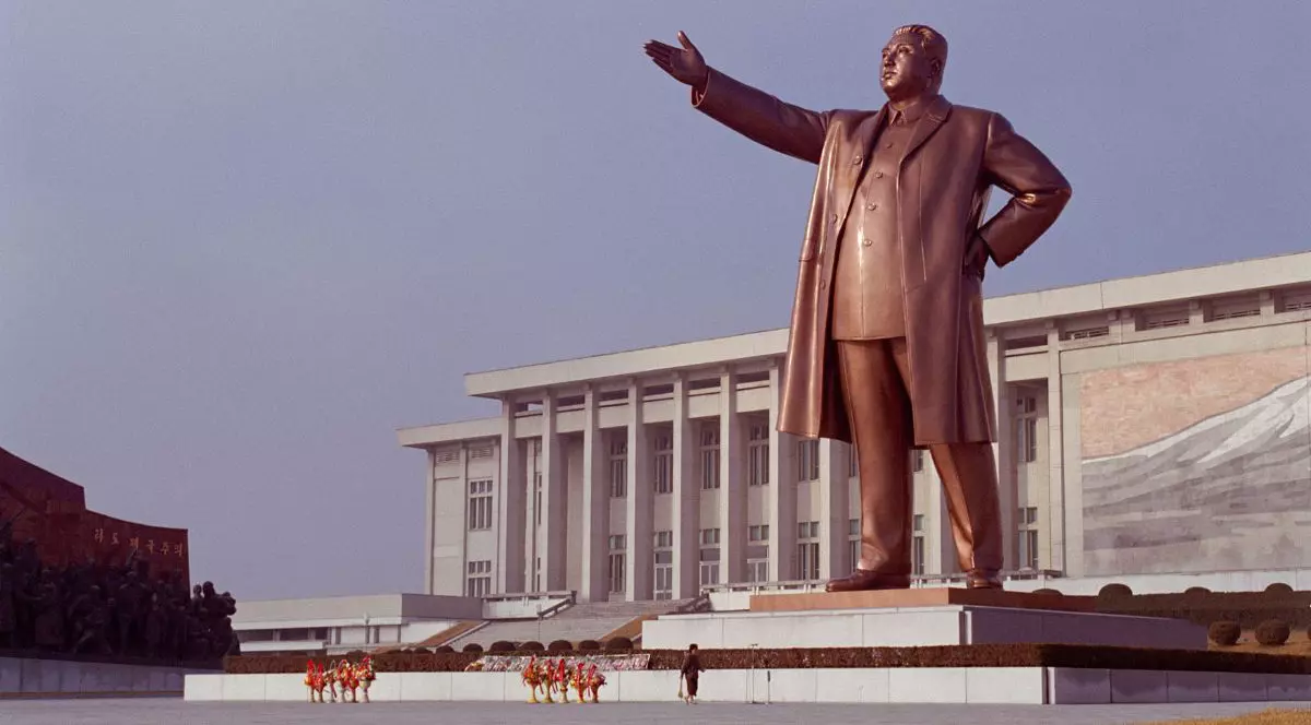 monument înalt, din material de culoarea bronzului, care îl înfățișează pe liderul nord-coreean Kim Jong-Un, cu o mână în șold și cealaltă mână întinsă în față, în fața unei clădiri albe cu coloane, Pyongyang, Coreea de Nord