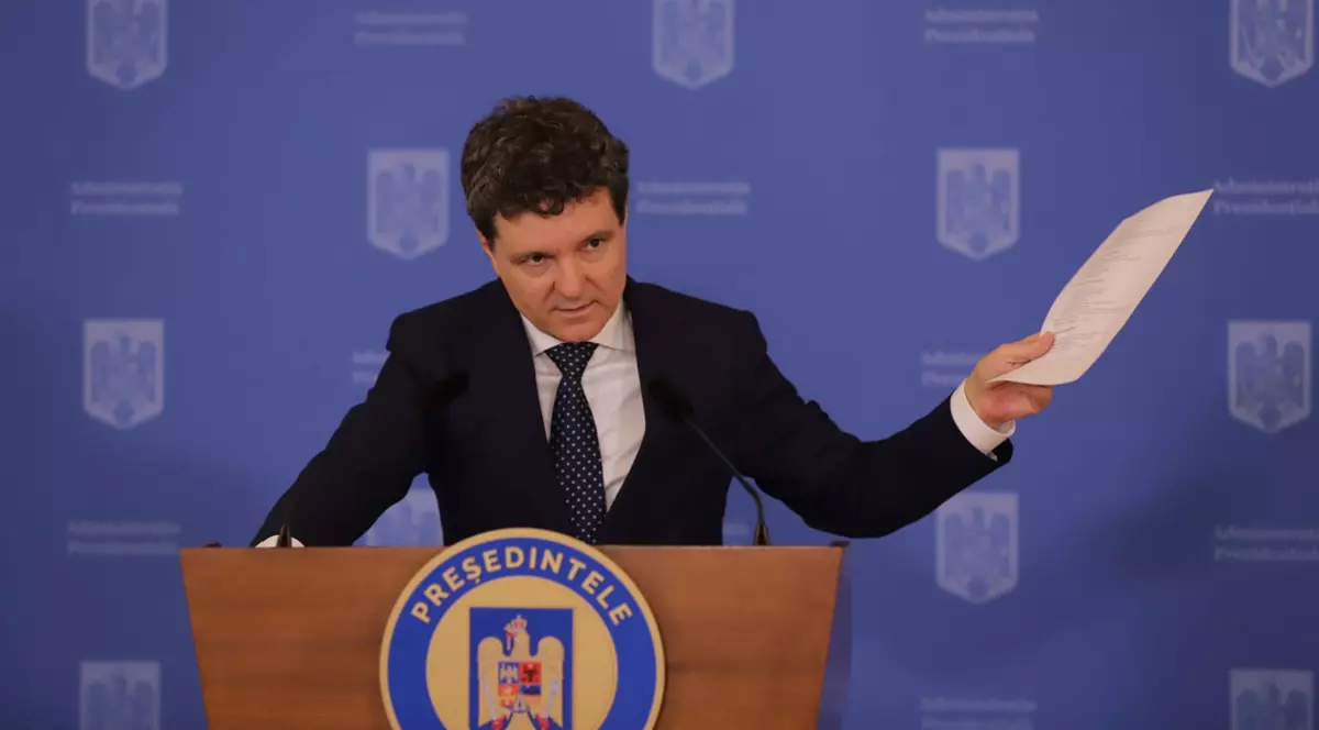 Nicușor Dan la pupitru, în conferința de presă din 14 iulie 2025, îmbrăcat într-un costum negru, cu o cămașă albă și o cravată neagră, având în mâna dreapta un document printat și ținând mâna stângă pe pupitru, cu capul ușor plecat