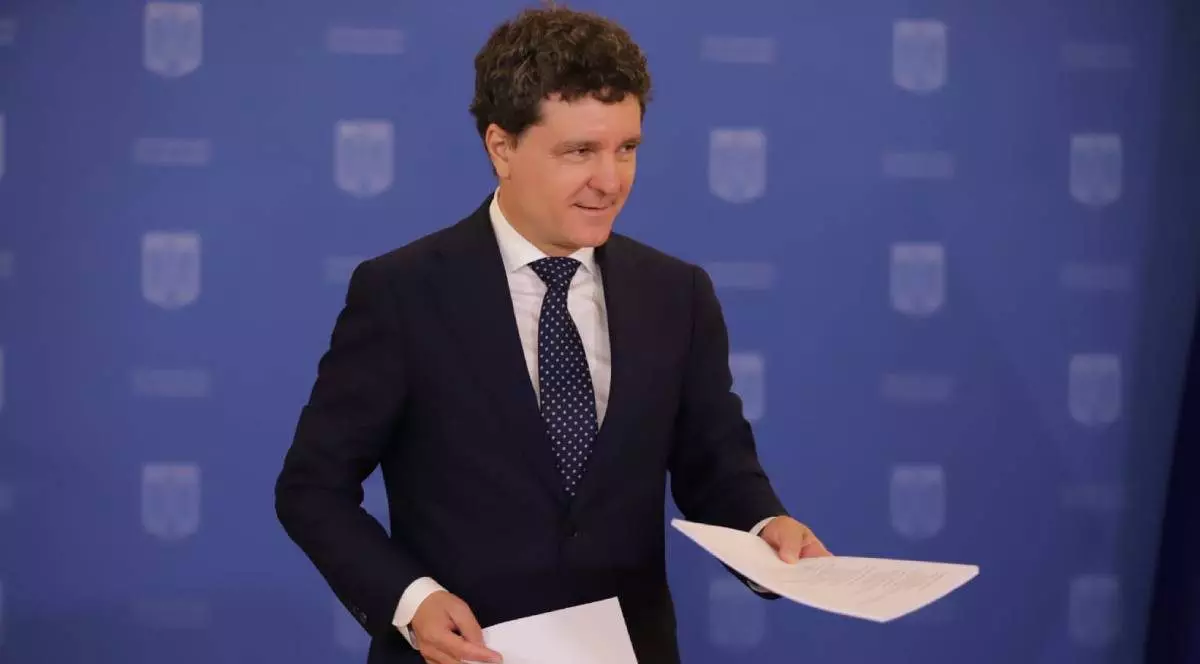 Nicușor Dan surprins în timpul conferinței de presă de luni, 14 iulie 2025, în timp ce ține în mâini hârtii. Poartă un sacou negru, o cămașă albă și o cravată albastră cu buline albe