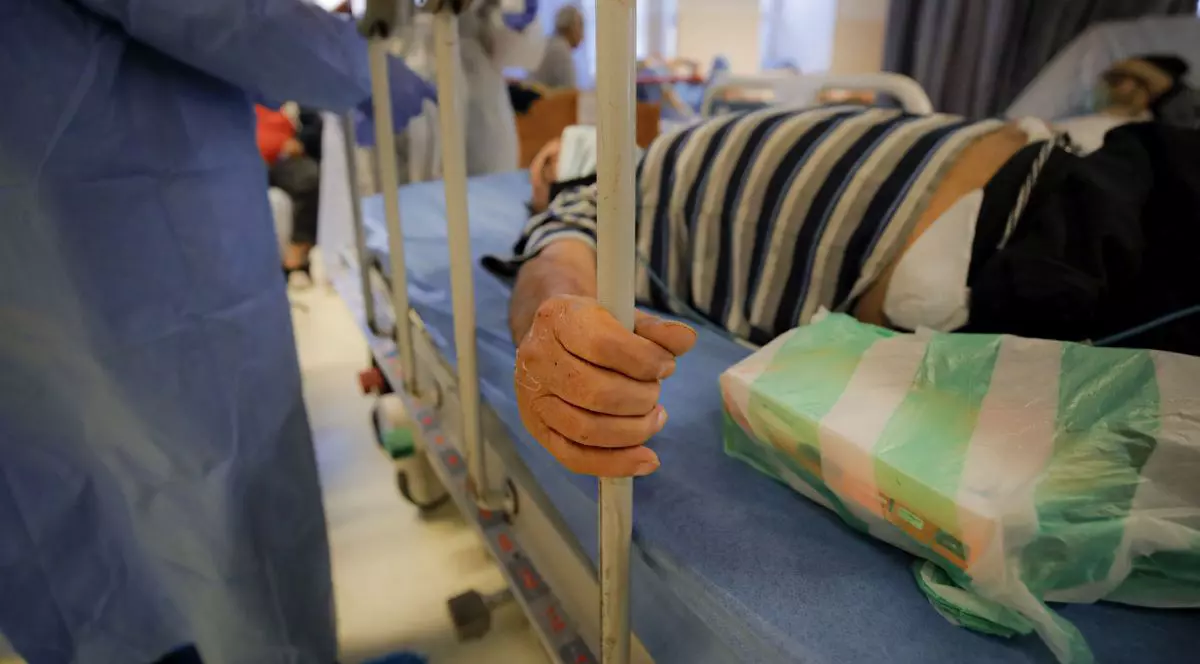Pacient pe un pat la Unitatea de Primiri Urgențe a unui spital din România, în timp ce se ține cu o mână de gratiile patului, iar lângă pat este un cadru medical îmbrăcat cu o uniforma albastră