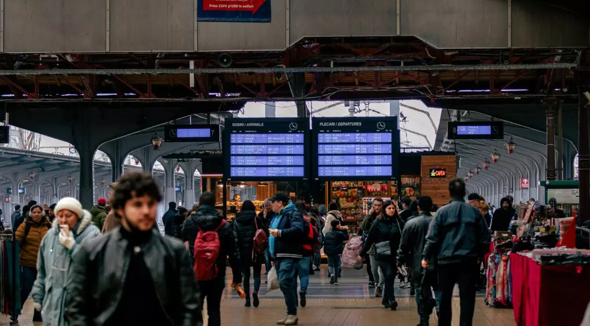 Imagine creativă cu Gara de Nord din București, în prim plan, în blur, mai mulți călători, iar pe fundal panourile de informare cu plecări și sosiri ale trenurilor