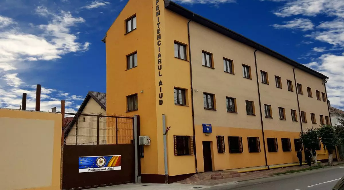 fațada clădirii penitenciarului de maximă securitate din aiud - clădire cu 2 etaje, vopsită în galben, pe care este scris în stil vertical „Penitenciarul Aiud” și o poartă metalică vopsită în maro, pe care este lipit un banner în culorile drapelului României, pe care apare textul „Penitenciarul Aiud” și stema Ministerului Justiției