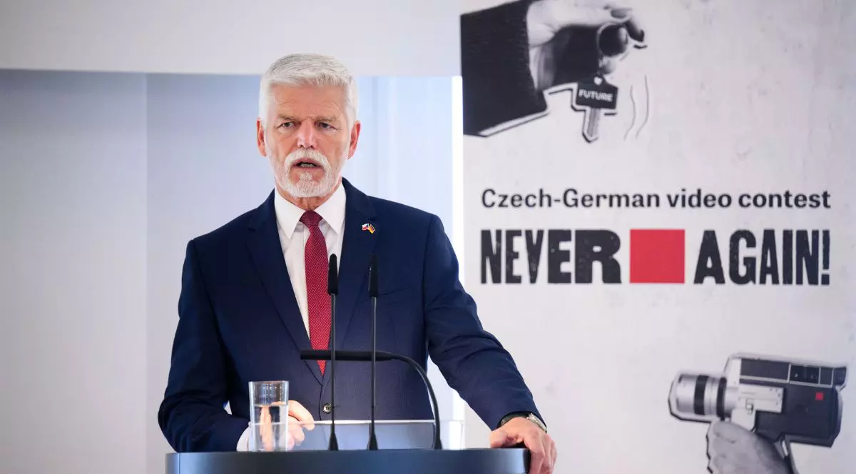 Petr Pavel, îmbrăcat într-un costum negru cu cămașă albă și o cravată roșie, vorbind în fața unui pupitru de lemn vopsit negru, la microfon. Mimica sa facială este serioasă, și ține o mână aproape de corp și o alta pe marginea pupitrului