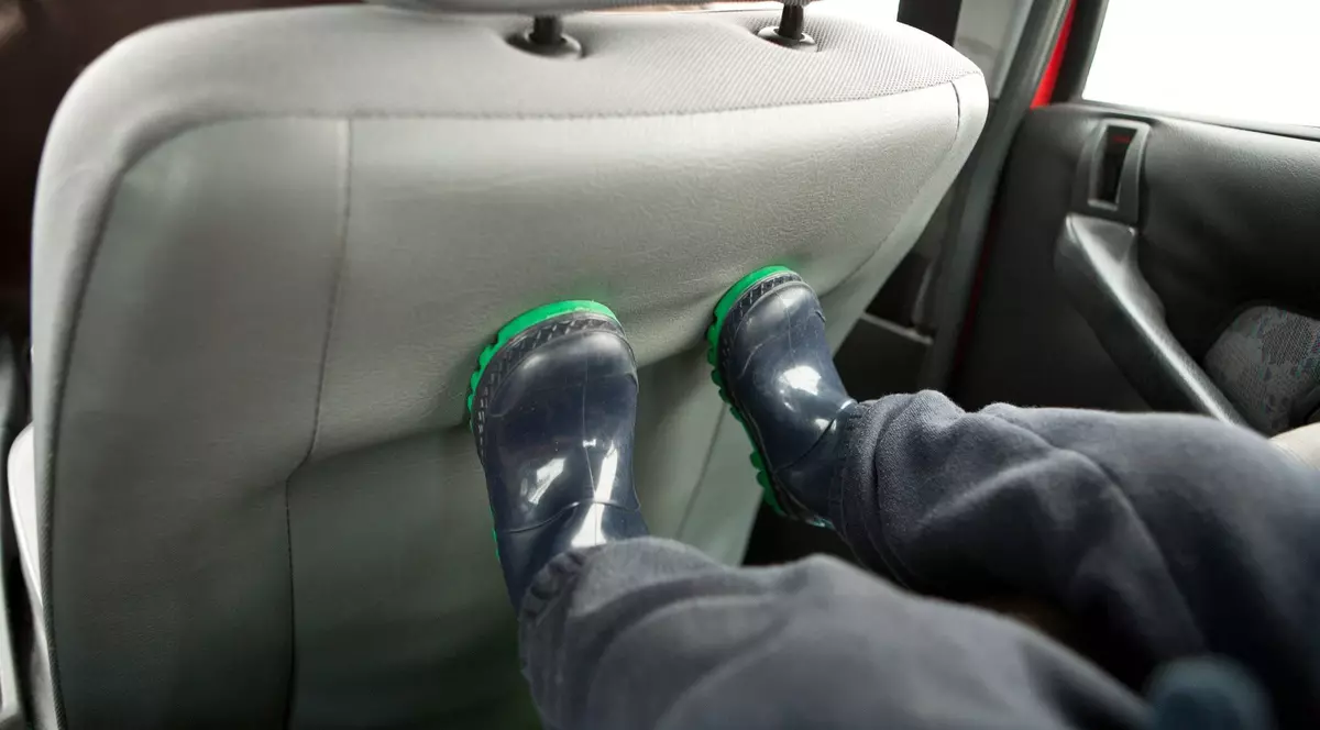 Imagine cu picioarele unui copil pe un scaun auto. Acesta poartă încălțăminte de culoare închisă și cu talpă verde și pantaloni negri