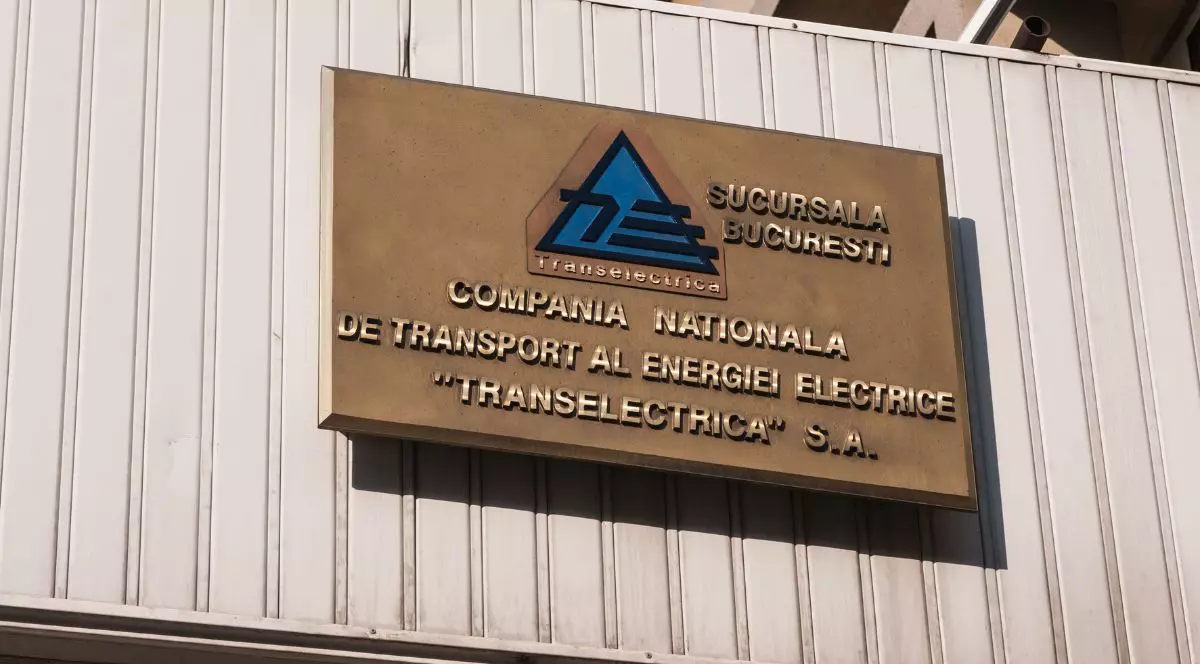 plăcuță metalică afișată pe fațada unei clădiri cu logo-ul companiei de stat Transelectrica și textul „Compania Națională de Transport al Energiei „Tranelectrica” S.A., scris cu litere metalice aurii pe un fundal maro