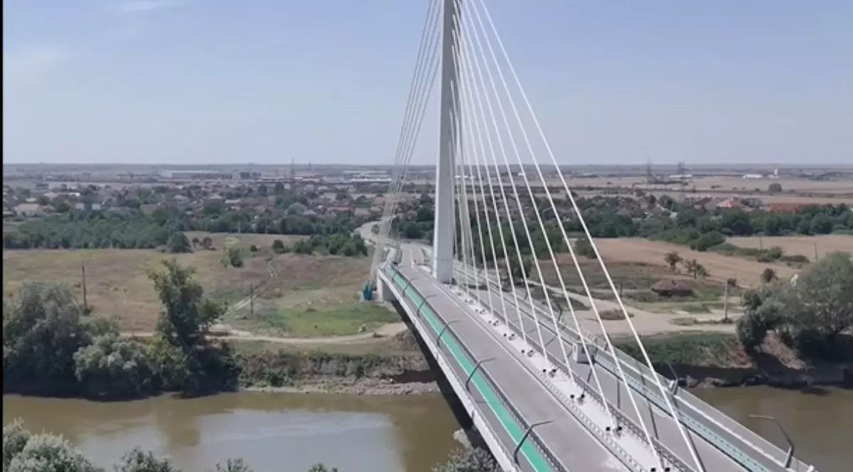 podul construit peste Mureș în județul Arad care traversează de la marginea inferioară a imaginii spre partea centrală-superioară. Podul este susținut de mai multe cabluri albe întinse oblic între un pilon vertical central și marginile podului, formând linii drepte și paralele. Lățimea podului permite traficul pe două benzi, separate de o zonă verde îngustă pe fiecare parte