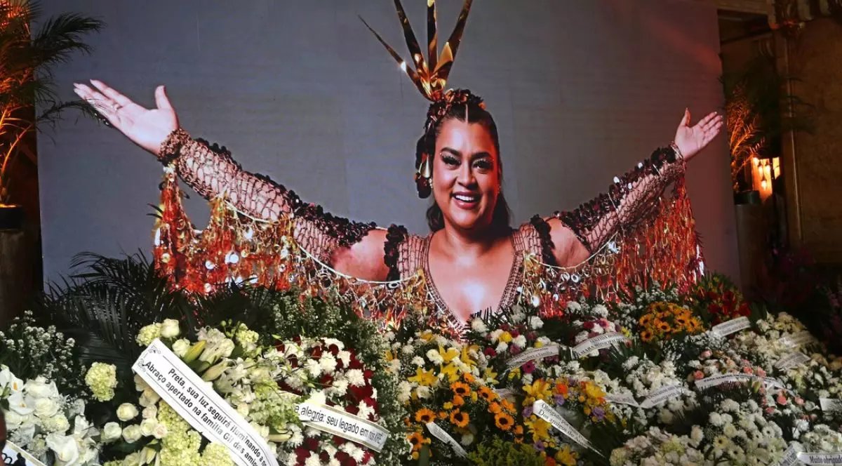 Imagine cu o fotografie a cântaretei braziliene Preta Gil, cu mainile deschise, inconjurată de flori, coroane și mesaje. Preta Gil, fiica lui Gilberto Gil, a murit pe 20 iulie 2025