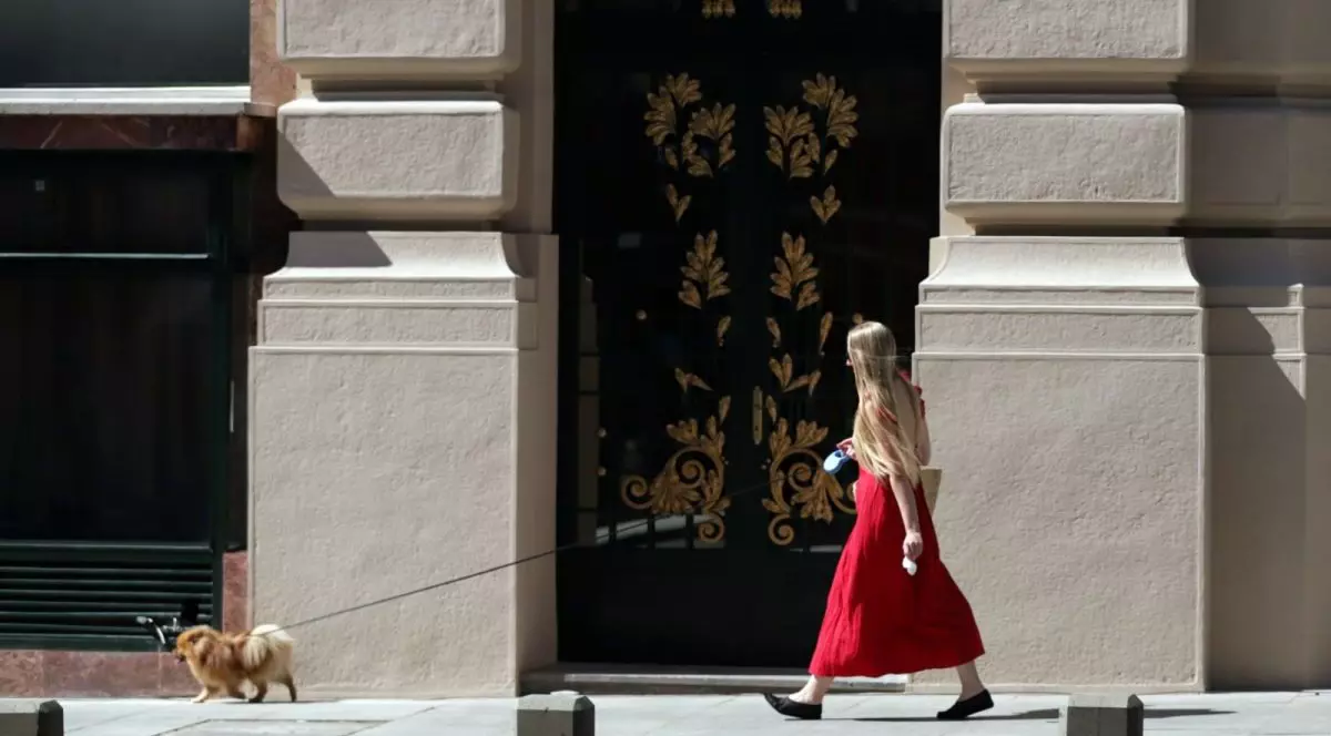 O tânără cu părul blond și o rochie roșie, lungă, își plimbă cățelul ținut în lesă și trece prin dreptul unei clădiri cu uși impozante, într-o zi de vară, 2 iulie 2025