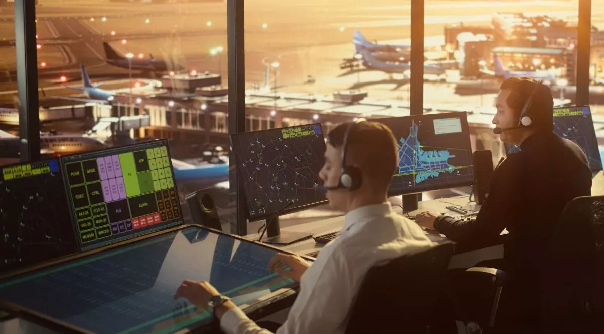 Echipă diversă de control al traficului aerian care lucrează într-un turn modern al aeroportului la apus. Biroul este plin de monitoare de computere desktop cu ecrane de navigație și date radar de zbor pentru controlori.