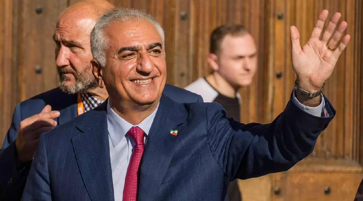 Reza Pahlavi face semn cu palma deschisă și zâmbește jurnaliștilor