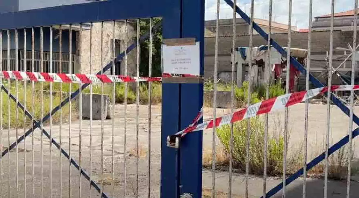 O poartă legată și securizată cu bandă a poliției, în spatele ei clădiri abandonate