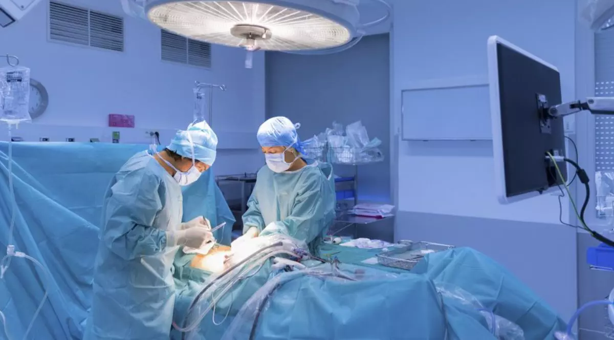 doi chirurgi in sala de operație, aplecati asupra unui pacinet, intr-o lumina albastra, cu o lapa mare deasupra mesei de operație.