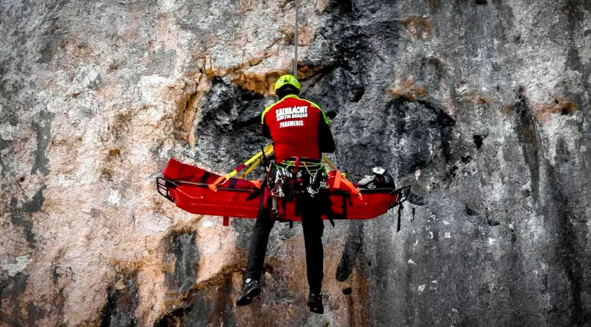 o persoană văzută din spate, îmbrăcată într-o vestă roșie și cască galbenă, suspendată cu ajutorul unor corzi în fața unei stânci verticale. Persoana stă lângă o targă rigidă roșie, prinsă de sistemul de prindere al corzilor. În fundal se vede suprafața aspră, gri și brună a stâncii. Pe vesta persoanei sunt inscripții albe cu textul „Salvamont Județul Brașov - Paramedic”