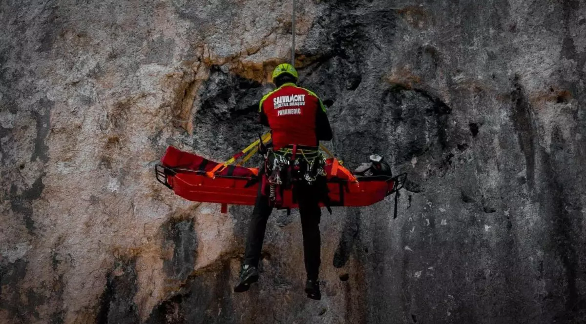 Un salvamontist din Brașov cu echipament roșu și cască de protecție îl coboară de pe o stâncă pe un bărbat care este pe o targă trasă cu coardă