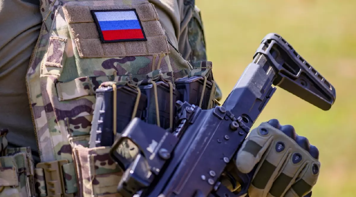 Imaginea arată un soldat îmbrăcat în uniformă de camuflaj cu steagul Rusiei pe vestă, ținând o armă automată în mână.