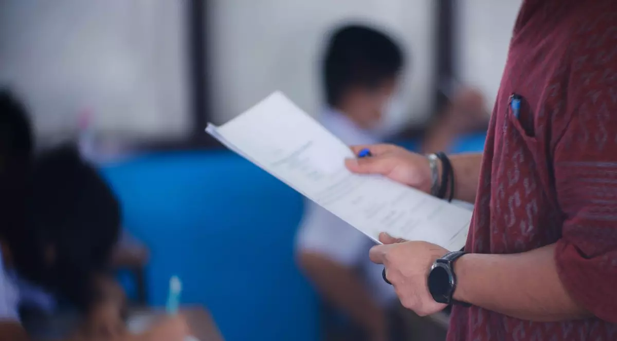 Imagine cu un tânăr care ține în mâini o foaie de examen. Poartă un tricou roșu închis și un ceas.