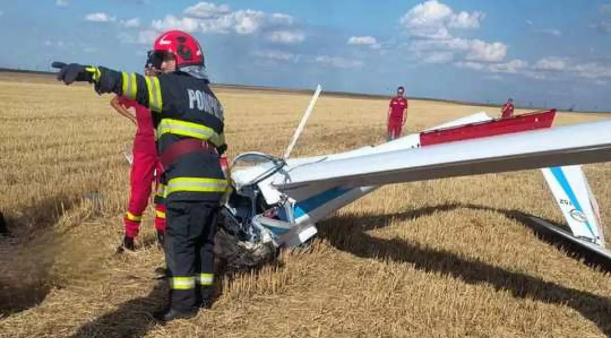 Fotografia arată o zonă deschisă, acoperită cu iarbă uscată sau paie, sub un cer albastru cu câțiva nori albi. În centrul imaginii se află resturile unui aparat de zbor de dimensiuni mici, cu aripa ruptă și fuzelajul avariat. În jurul epavei se află mai multe persoane. Una dintre ele, îmbrăcată într-o uniformă de pompier neagră cu dungi reflectorizante galbene și cuvântul „POMPIERI” pe spate, poartă cască roșie și arată cu mâna într-o direcție. Alte persoane poartă uniforme roșii ale SMURD