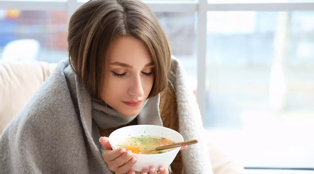 Tânără bolnavă, învelită cu o pătură gri, în timp ce mănâncă dintr-o farfurie cu supă, pe pat, lângă un geam